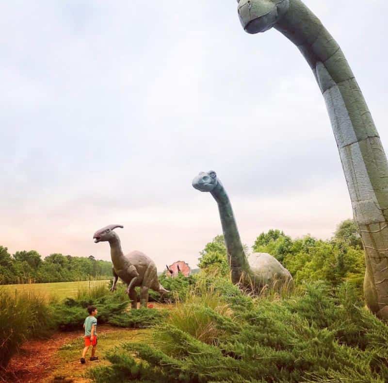 A child in a red shirt stands in front of large dinosaur sculptures in a grassy area, with trees and a cloudy sky in the background.