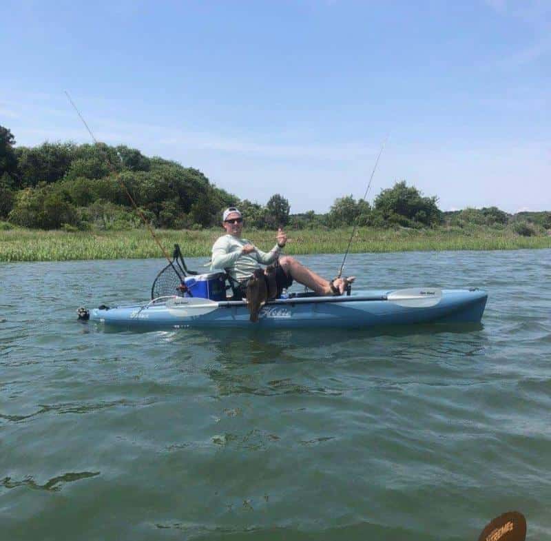 Person in a blue kayak on a lake, holding a fishing rod and a fish. Trees and grass are visible in the background under a clear sky.