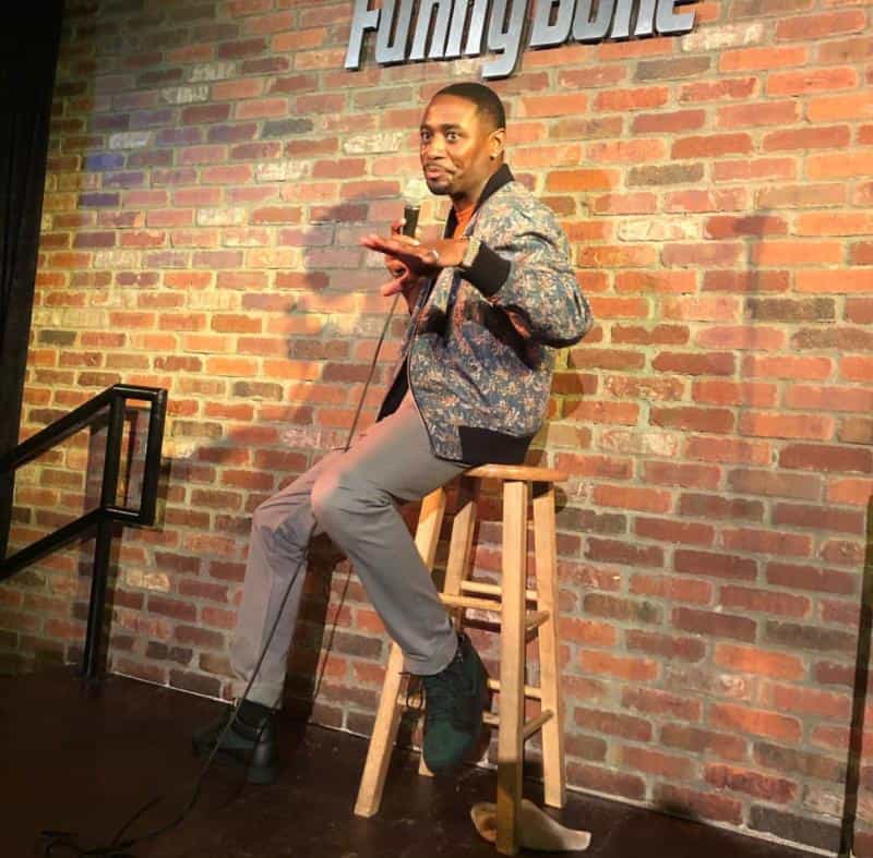 Comedian performs on stage at a comedy club, holding a microphone and gesturing, with a brick wall background.