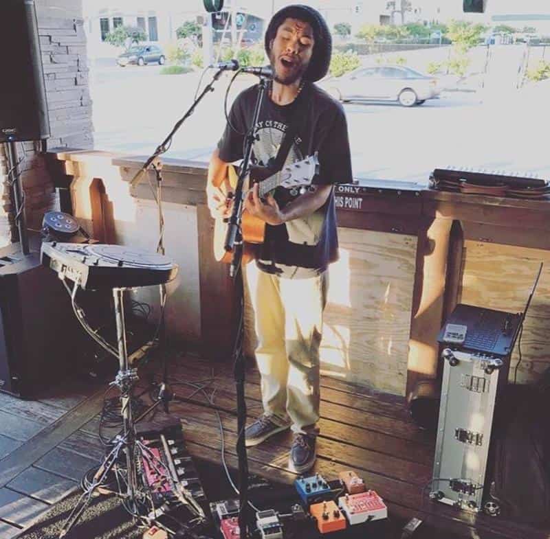 Musician playing guitar and singing into a microphone on a small stage, with various audio equipment and pedals at his feet.
