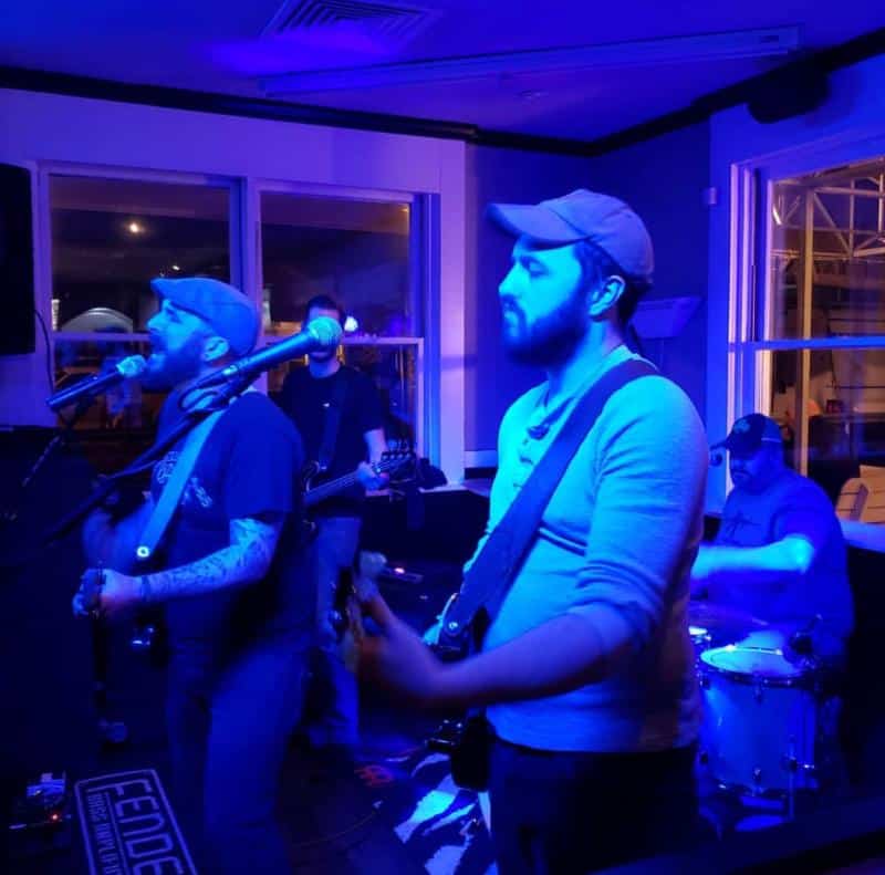A band performs live on stage under blue lighting. The guitarist sings into a microphone while the drummer plays in the background.