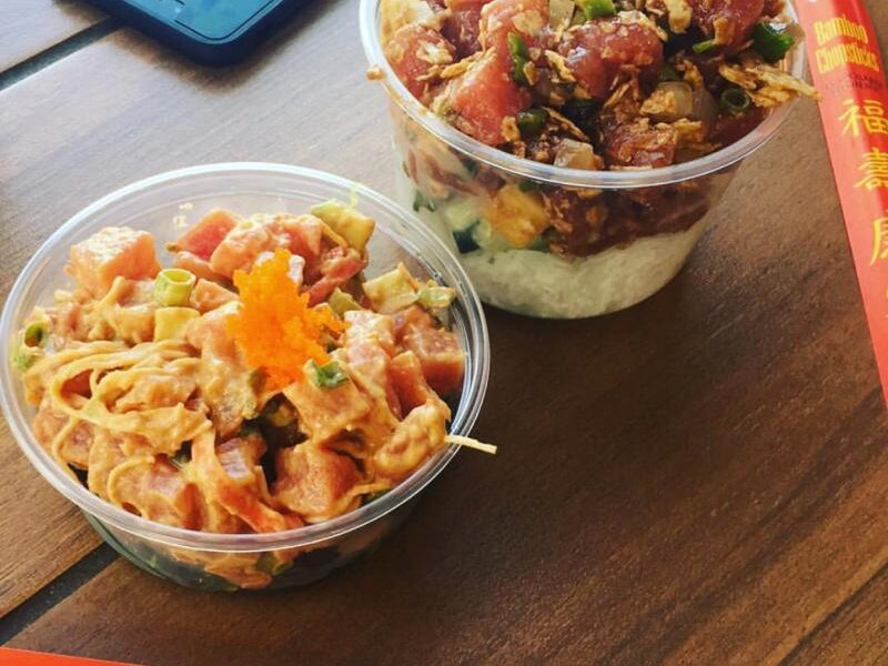 Two bowls of poké on a wooden table. One has a spicy mayo topping and the other has a soy-based sauce. Red chopsticks and a smartphone are nearby.