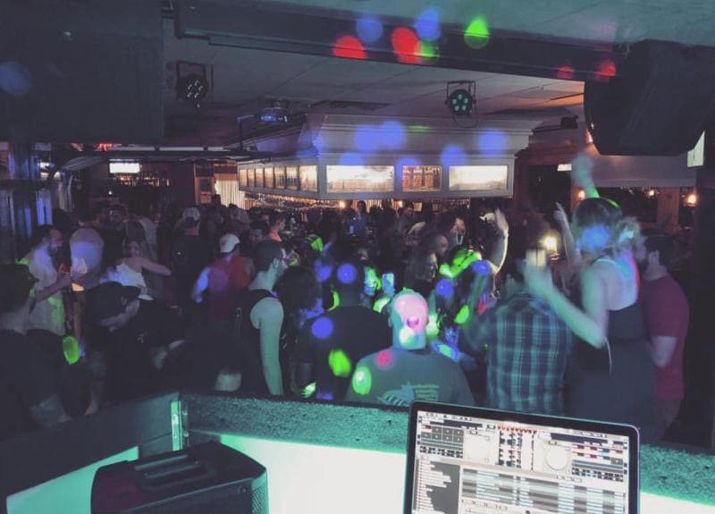 A crowded nightclub with colorful lights and people dancing. A DJ booth is visible in the foreground with DJ equipment.