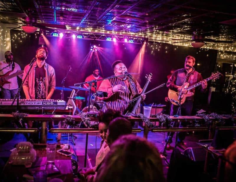 A band performs energetically on stage with colorful lights in the background. A keyboardist, singer, and guitarist are visible. Audience members watch from tables in the foreground.