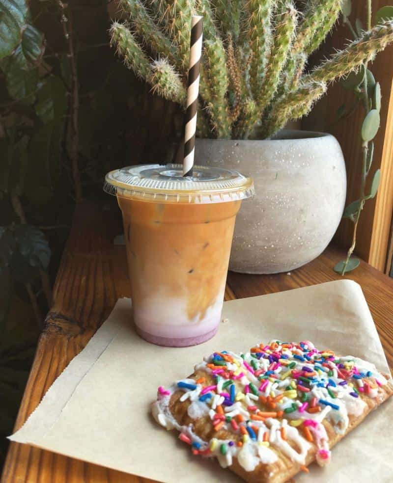 Iced layered drink with a striped straw next to a pastry topped with colorful sprinkles on brown paper. Potted cactus in the background.