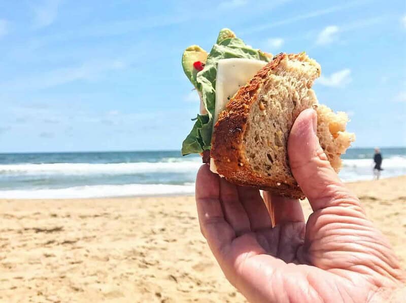 A hand holds a sandwich with lettuce, cheese, and tomato, set against a sandy beach and ocean backdrop under a blue sky.