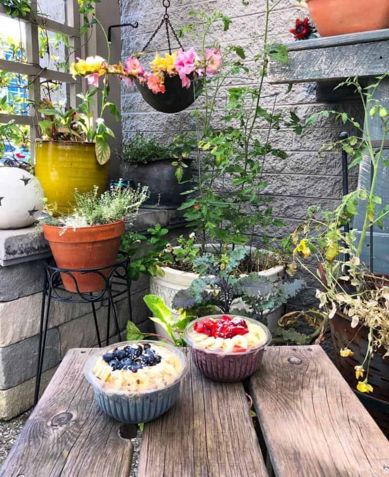 Two fruit-topped smoothie bowls on a wooden table, surrounded by potted plants and flowers on a rustic patio.