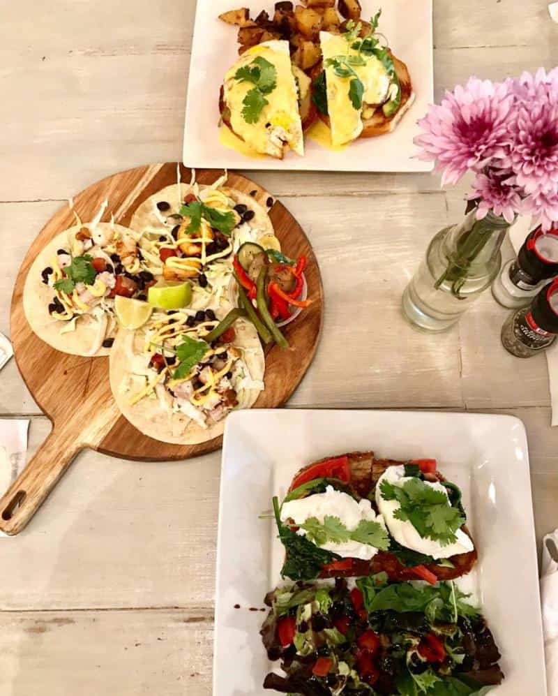 Two plates of food with tortillas on a wooden board, a dish with eggs and potatoes, and flowers in a vase on a table.