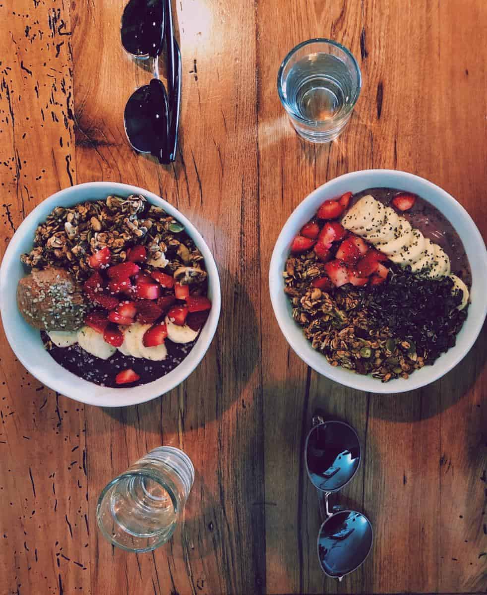 Two bowls of yogurt topped with granola, sliced bananas, strawberries, and chia seeds on a wooden table. Two pairs of sunglasses and two glasses of water are beside the bowls.