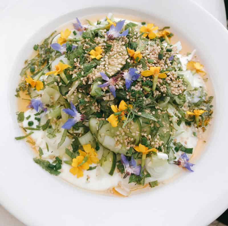 A white plate with a salad topped with cucumber slices, herbs, sesame seeds, and edible flowers, all on a creamy dressing.