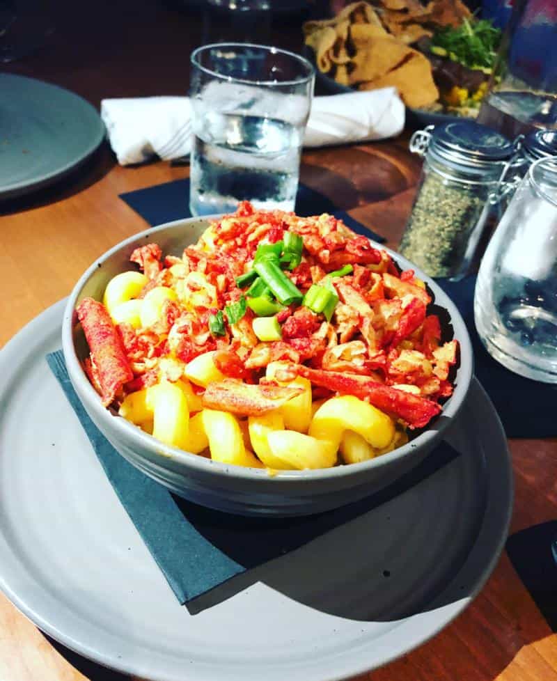 A bowl of pasta topped with chopped red pepper, green onions, and crumbled sausage. A glass of water and seasoning bottles are on the wooden table.