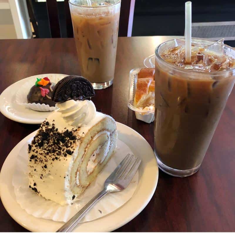 A table with iced coffees, a slice of cake topped with an Oreo, a decorated cake pop, and another dessert in a tray.