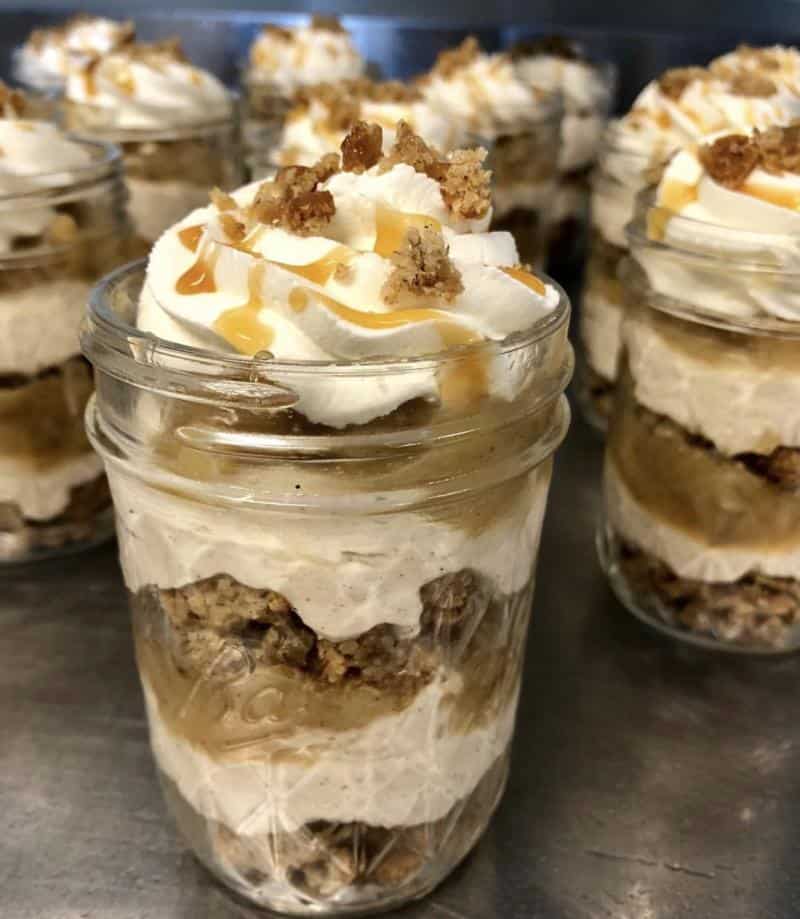 Several dessert jars filled with layers of whipped cream, caramel, and crumbled topping, displayed on a metal surface.