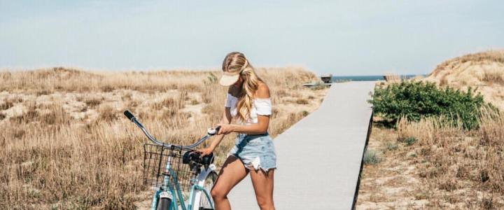 A woman in a white top and denim shorts stands beside a bicycle on a sandy path with dunes in the background.