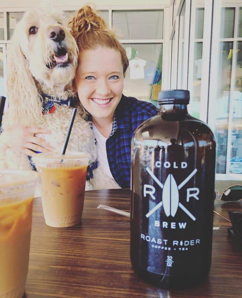 Woman smiling with a dog, sitting at a table with iced coffee and a bottle labeled "Cold Brew Roast Rider.