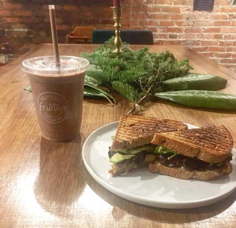 A grilled sandwich with avocado and greens on a plate beside a chocolate smoothie in a cup with a straw, set on a wooden table with greenery and a brick wall backdrop.