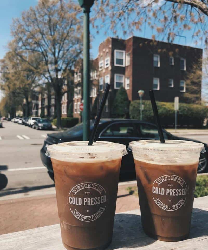 Two plastic cups of iced coffee with black straws on a wooden bench, in front of a street with parked cars and brick buildings, under a clear sky.