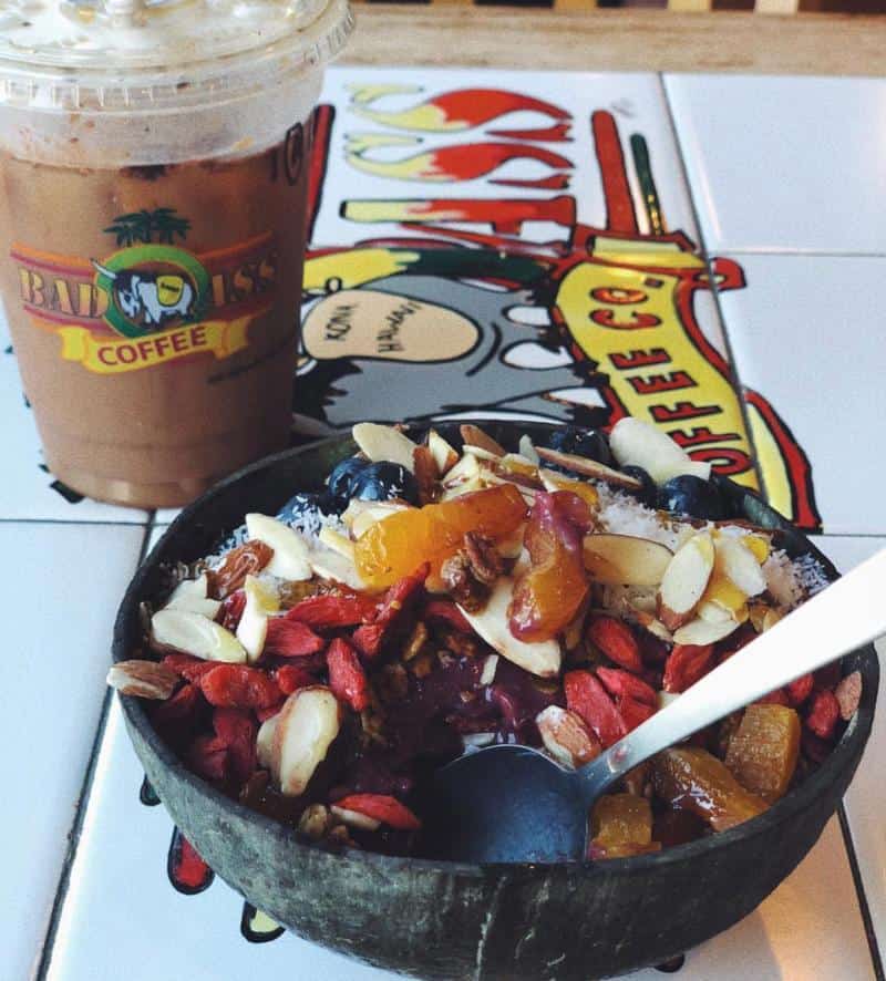Bowl of mixed fruit, nuts, and seeds with a spoon, next to an iced coffee on a tiled table.