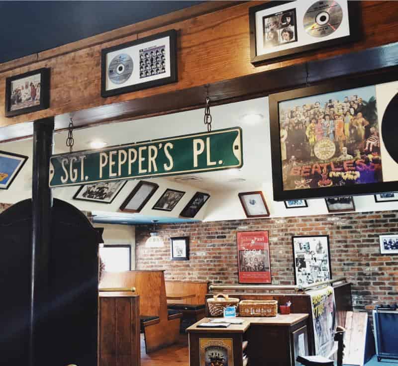 A restaurant interior with Beatles memorabilia, including a "Sgt. Pepper's Pl." sign, album covers, and framed photos on brick and wood walls. Booth seating is visible.