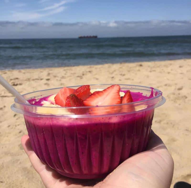 A hand holds a bowl of vibrant smoothie topped with sliced strawberries and banana against a sandy beach and ocean backdrop.