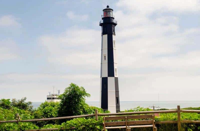 Black and white lighthouse with a two-tiered top, surrounded by greenery, under a partly cloudy sky.