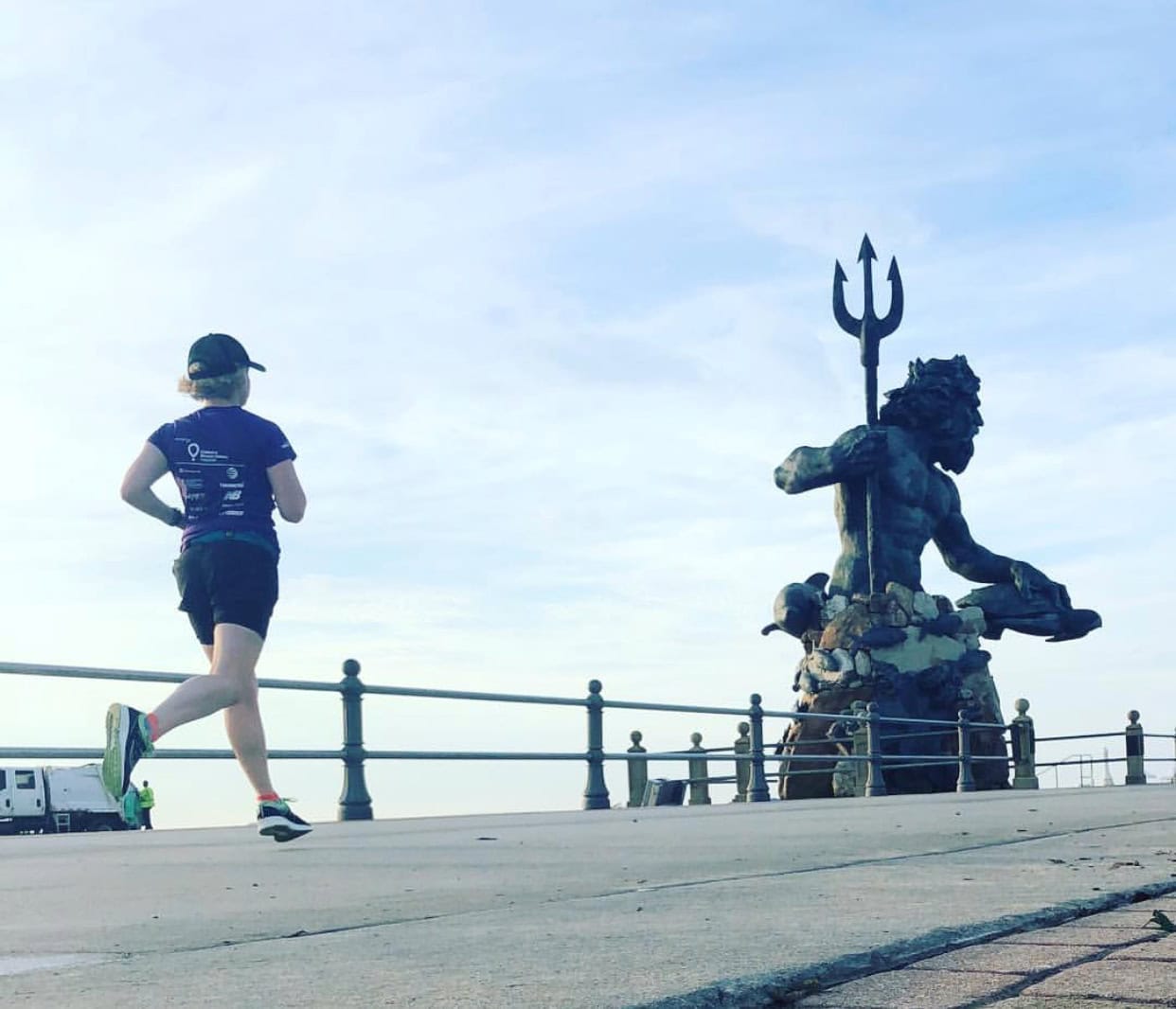 Runner on a seaside boardwalk passing a large statue of Neptune holding a trident.
