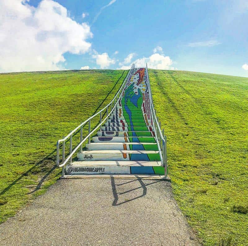Wide grassy hill with stairs featuring colorful mural art of a person climbing, under a blue sky with clouds.