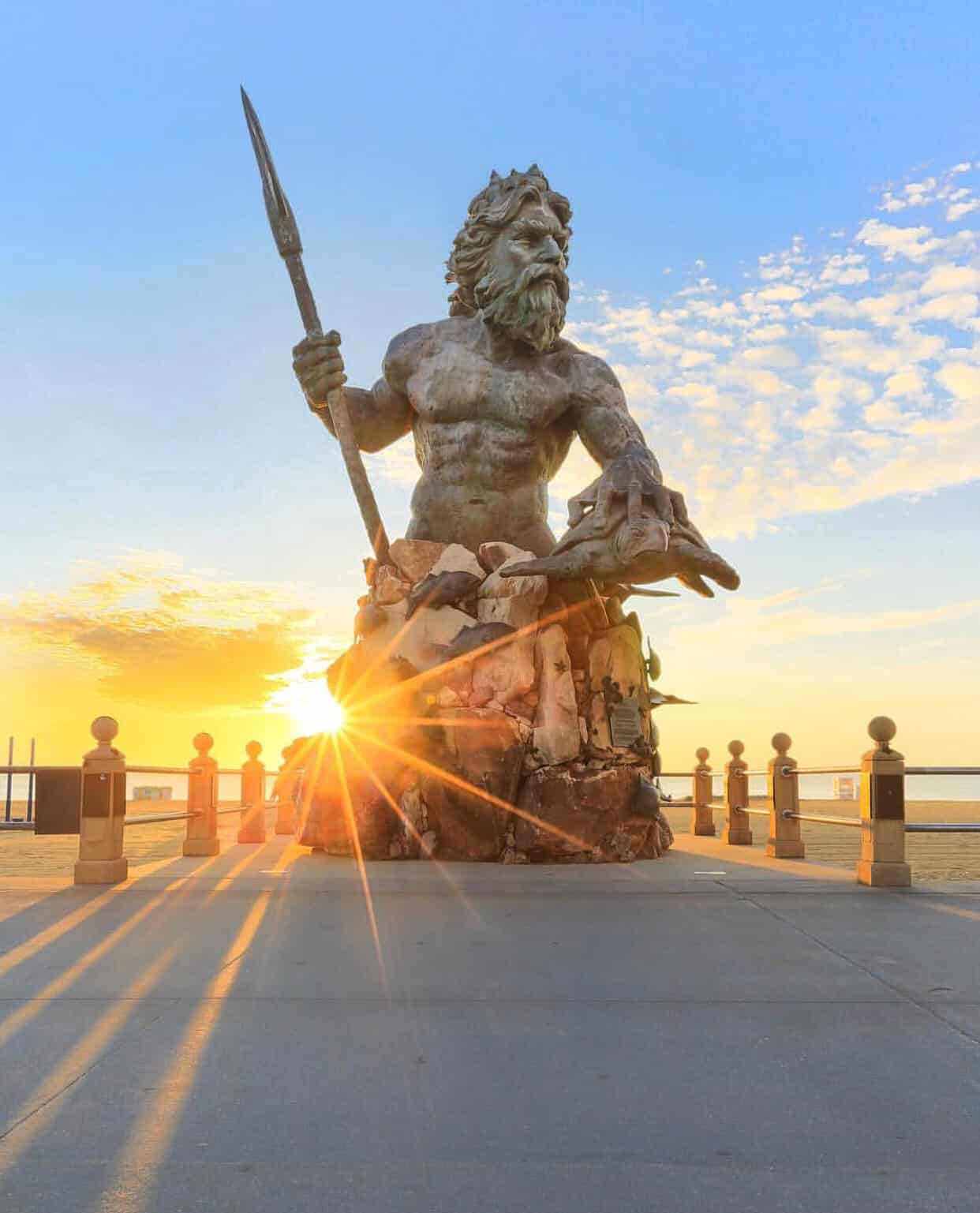A large statue of a bearded man holding a trident with the sun setting behind it, creating a starburst effect. The statue sits on a beachfront promenade with clear skies and scattered clouds.