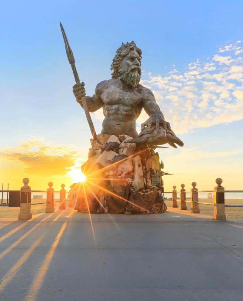 A large statue of a bearded man holding a trident is silhouetted by the sunrise on a boardwalk.