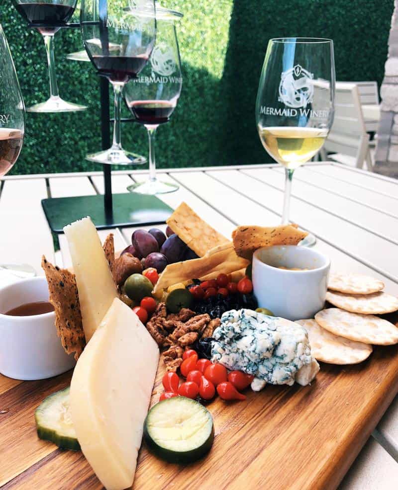Cheese and charcuterie board with assorted cheeses, crackers, nuts, grapes, olives, sliced cucumber, and red berries, accompanied by glasses of wine on an outdoor table.