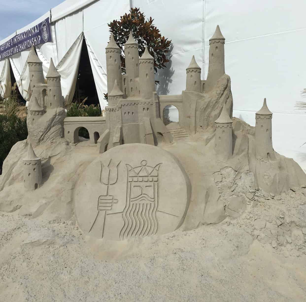 Elaborate sandcastle sculpture featuring multiple towers and a large etched emblem of a crowned figure holding a trident, displayed near a white tent.