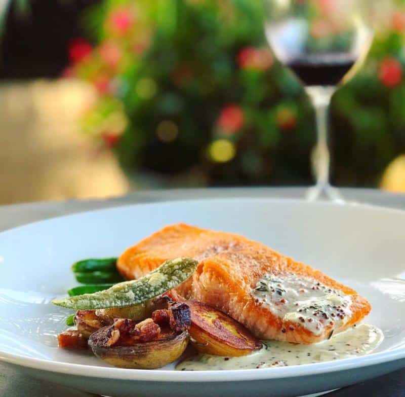 A plate of salmon with creamy sauce, accompanied by potatoes, green beans, and a wine glass in the background.