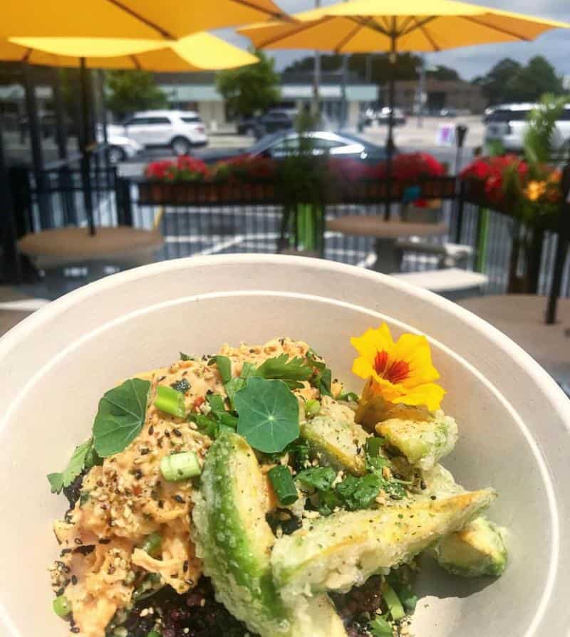 A bowl of quinoa topped with sliced avocado, spicy cream sauce, and edible flowers is set against an outdoor patio with yellow umbrellas and a parking lot in the background.