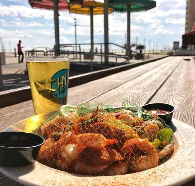 Plate of seasoned shrimp covered with a net, two small sauce containers, and a glass of beer on a wooden table, with a beachside view in the background.