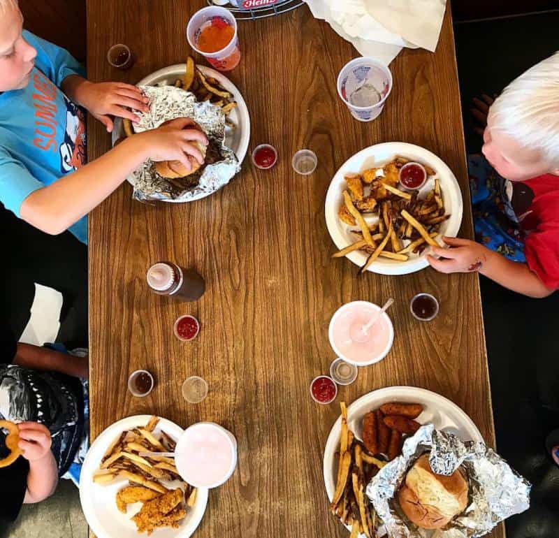 Four children sitting at a table eating burgers, fries, and chicken tenders with cups of soda. Plates are accompanied by small sauce cups.