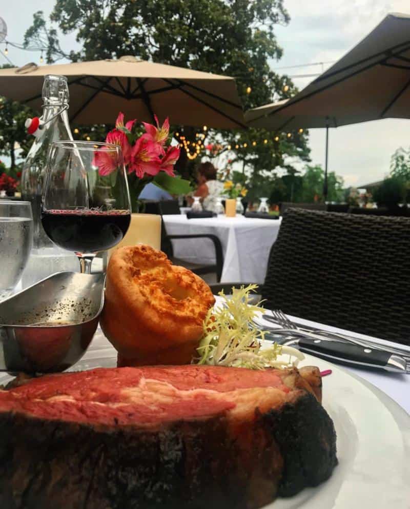 Plate with a cut of roast beef, Yorkshire pudding, and greens; accompanied by a glass of red wine. Outdoor dining setting with tables, chairs, and umbrellas in the background.