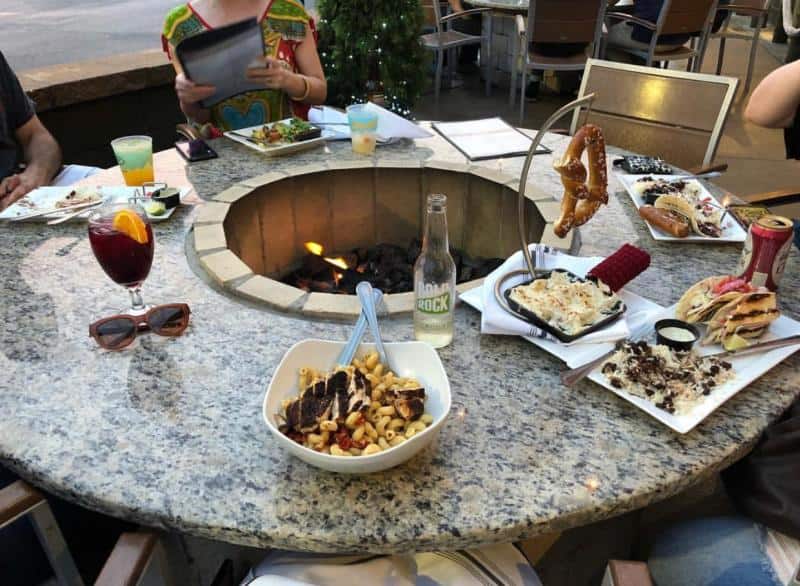 A variety of dishes and drinks are arranged on a circular stone table with a fire pit in the center. The table includes pasta, pretzels, and beverages.