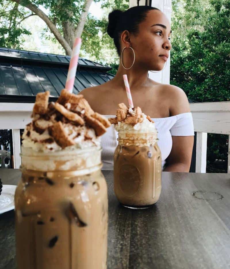 Woman sitting at a table outside with two iced coffee drinks topped with whipped cream and pastry pieces.