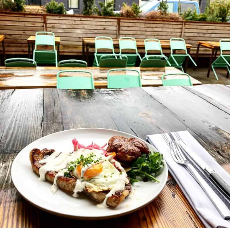 A plate with meat, an egg, white sauce, greens, and pickled onions on a wooden table. Empty green chairs are visible in the background.