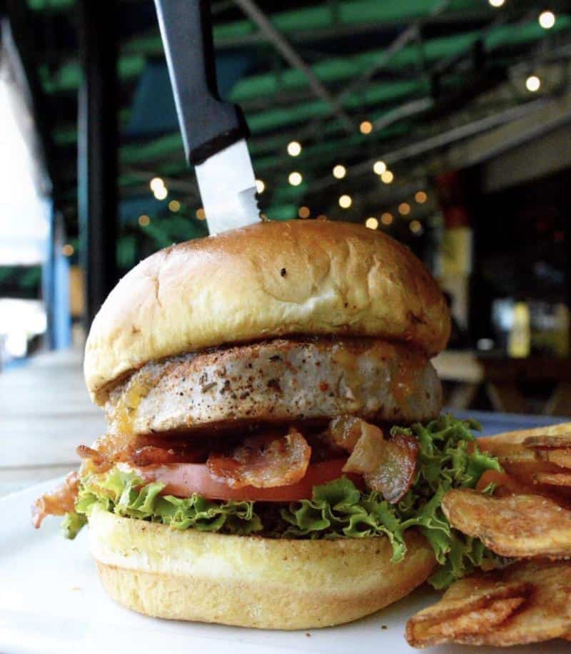 Burger with lettuce, tomato, bacon, and meat patty, topped with a knife, served with crispy potato chips on a white plate.