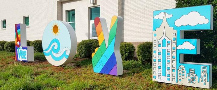 Colorful outdoor sculpture spelling "LOVE" with ocean, rainbow, and city designs on each letter, set against a white brick building and greenery.