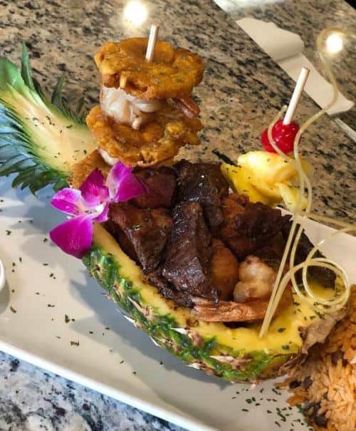 Pineapple boat filled with cooked meat and shrimp, garnished with an orchid and skewered fruit, served with a side of rice on a white plate.