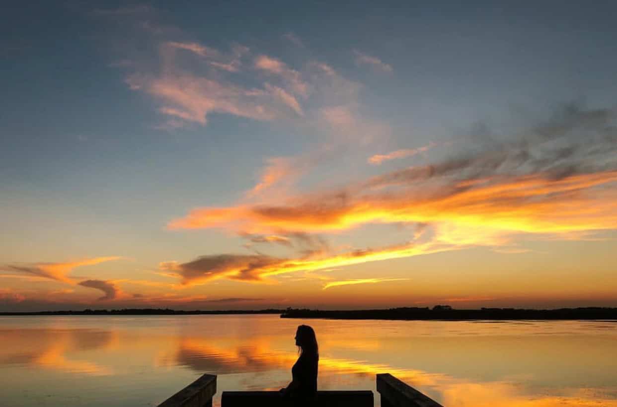 Silhouette of a person on a dock overlooking a calm lake at sunset, with vibrant orange and yellow clouds in the sky.