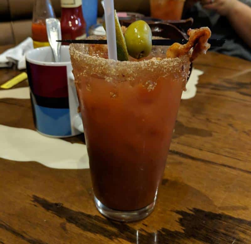 A glass of Bloody Mary with a salted rim, garnished with a lime wedge, olive, and bacon strip, sits on a wooden table.