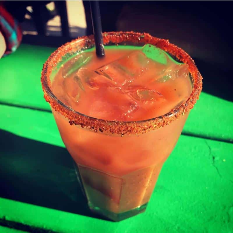 Iced beverage in a glass with a chili powder rim and a straw, placed on a green surface.