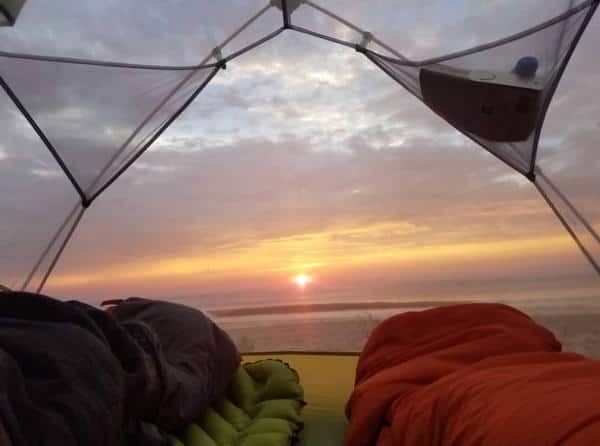 View from inside a tent showing a colorful sunrise over the horizon, with sleeping bags and gear visible in the foreground.