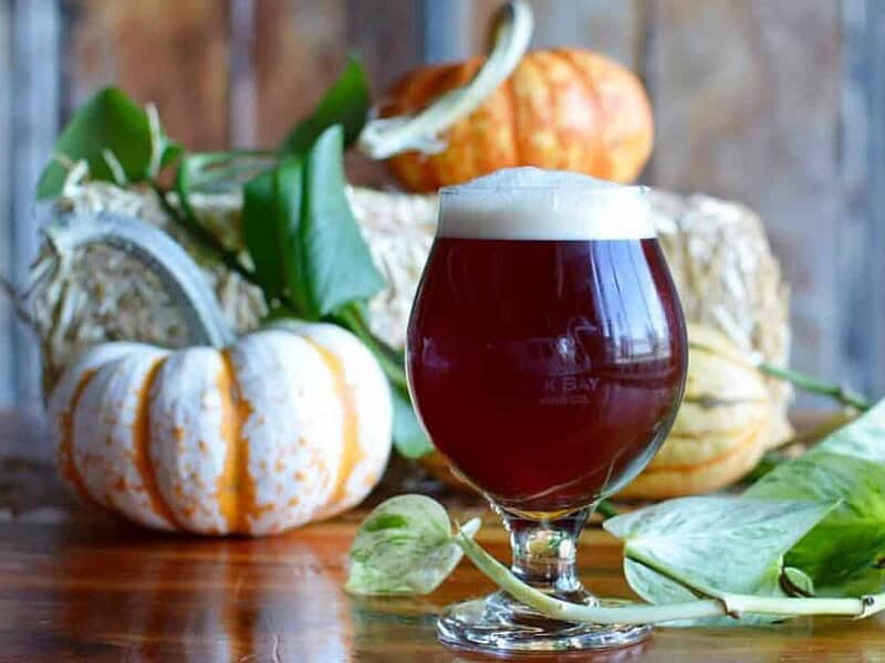 A glass of dark beer with a frothy head on a wooden table, surrounded by pumpkins and green leaves in the background.