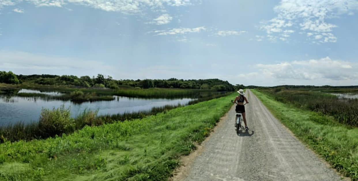 A person rides a bike on a gravel path between a grassy area and a body of water under a partly cloudy sky.