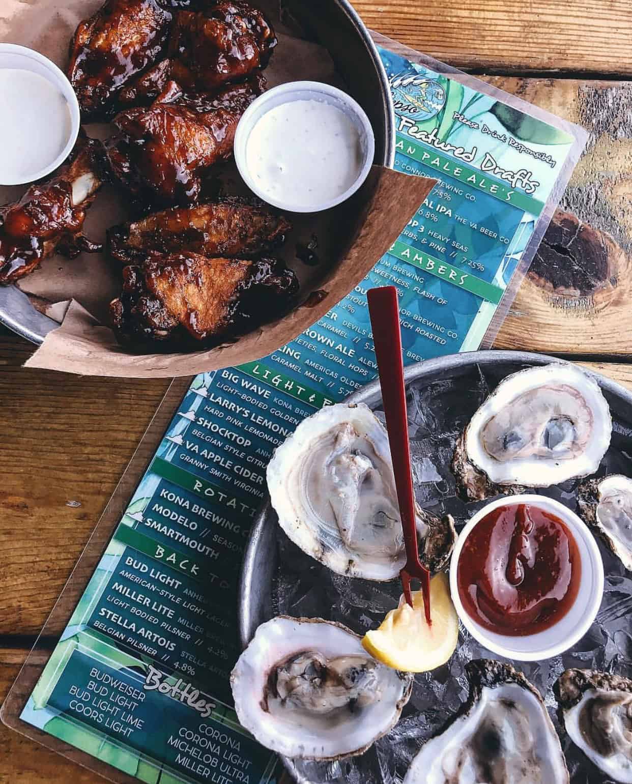 A table with grilled chicken wings, dipping sauces, and a platter of oysters with lemon slices and cocktail sauce, placed on menus.
