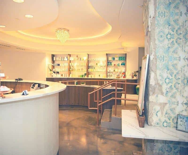 Modern reception area with a curved desk, warm lighting, and shelves displaying products. A chandelier hangs from the ceiling, and patterned wallpaper decorates the wall.